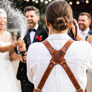 May include: A brown leather camera harness featuring an X-shaped back plate with metal rivets, adjustable straps, and metallic buckles. The harness is worn over a white shirt, designed to secure camera equipment across the shoulders and chest.