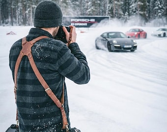 May include: A photographer wearing a grey knit beanie, a dark grey and black striped flannel shirt, and blue jeans, equipped with a brown leather dual camera harness, holds a camera to capture grey, red, and white sports cars on a snowy track.