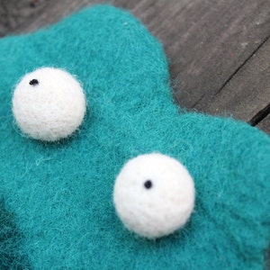 May include: A close-up of a handmade felt creature with two white, spherical eyes with black pupils. The creature is teal and has a textured, fuzzy appearance. The background is a wooden surface.