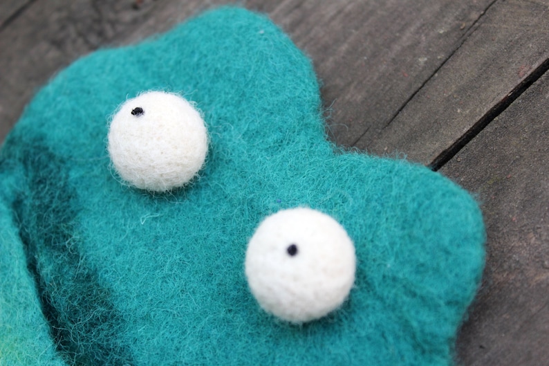 May include: A close-up of a handmade felt creature with two white, spherical eyes with black pupils. The creature is teal and has a textured, fuzzy appearance. The background is a wooden surface.