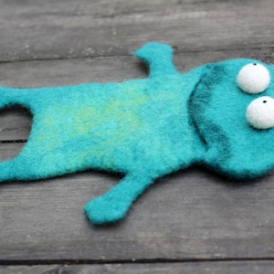 May include: A handmade teal felt frog with white, spherical eyes and a dark green outline. The frog is lying on a weathered wooden surface. The felt has a slight yellow-green marbling.