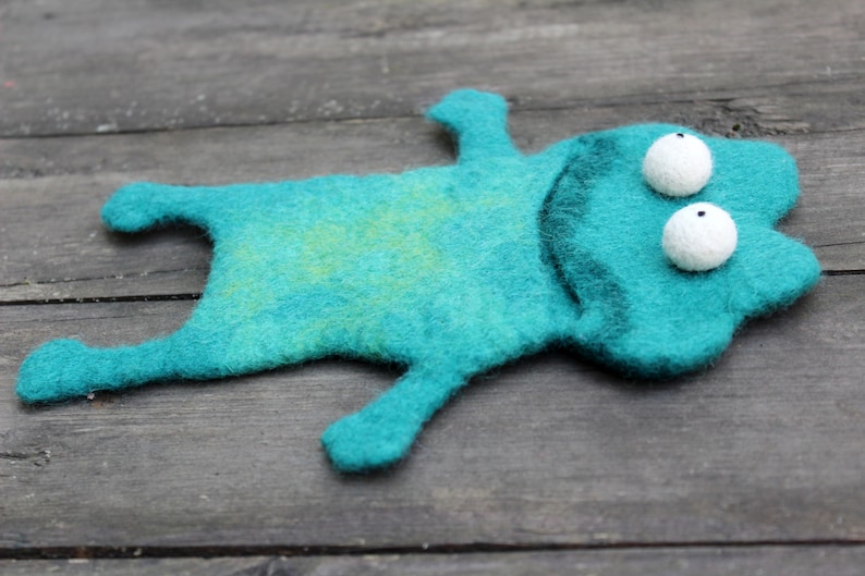 May include: A handmade teal felt frog with white, spherical eyes and a dark green outline. The frog is lying on a weathered wooden surface. The felt has a slight yellow-green marbling.
