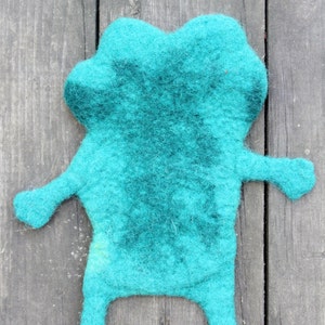 May include: A handmade teal felt frog-shaped item with a darker teal center. The frog has outstretched arms and legs, and is displayed on a weathered wooden surface. This whimsical item could be a toy or decorative piece.