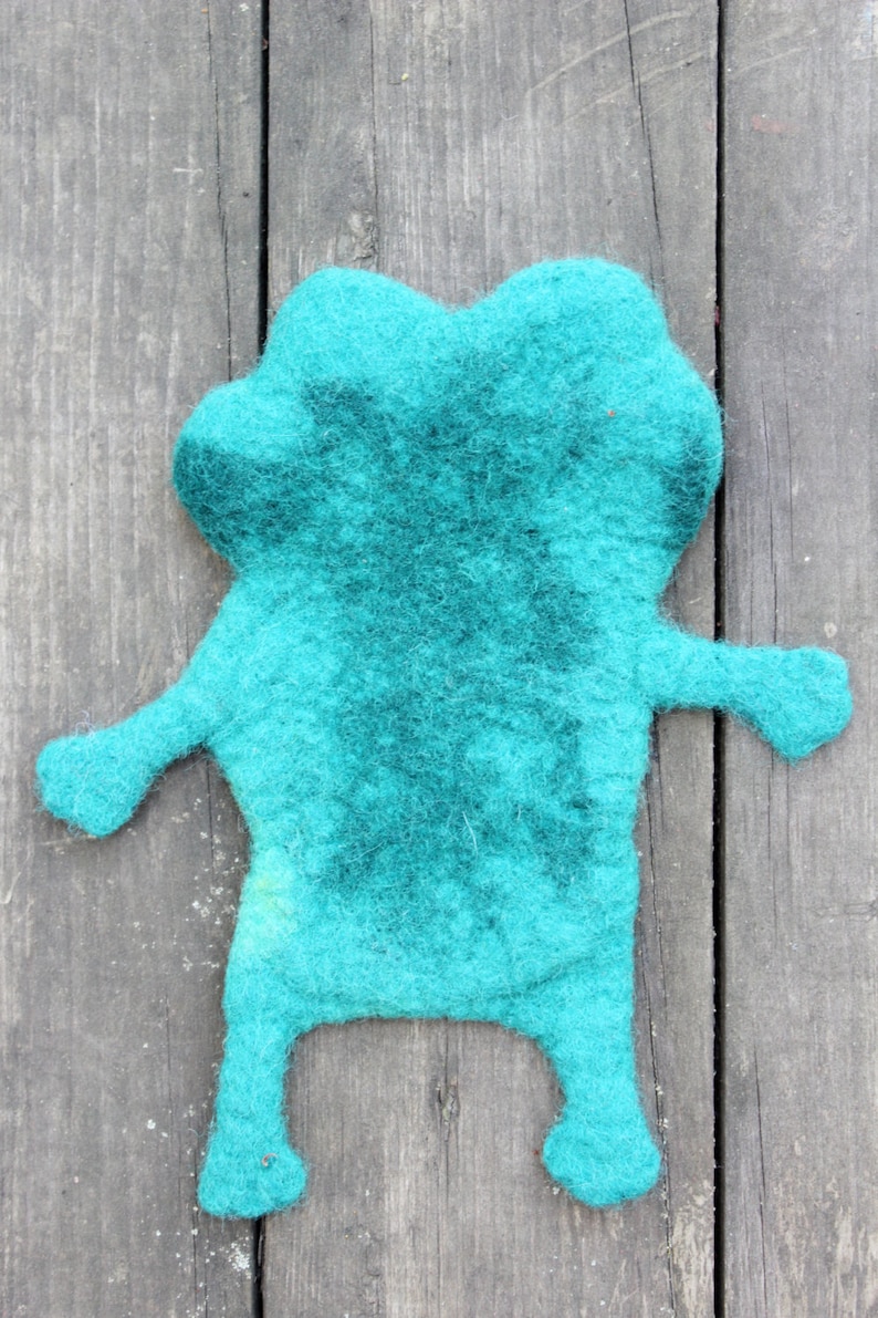 May include: A handmade teal felt frog-shaped item with a darker teal center. The frog has outstretched arms and legs, and is displayed on a weathered wooden surface. This whimsical item could be a toy or decorative piece.