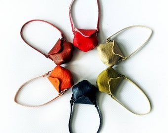 May include: A collection of small, handcrafted leather purses in a variety of colors, including red, brown, orange, and green. Each purse has a small handle and a unique, folded design. The purses are arranged in a circular pattern.
