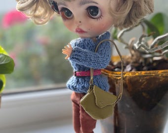 May include: A doll with large eyes and a vintage aesthetic, wearing a blue knit sweater, rust-colored pants, and a small olive green purse. The doll has a patterned headband and is posed near a window.