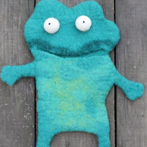 May include: A teal felt frog-shaped toy with white, round eyes and small arms and legs. The frog has a lighter green patch on its belly. The toy is handmade and sits on a weathered wooden surface.