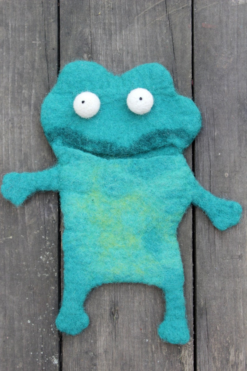 May include: A teal felt frog-shaped toy with white, round eyes and small arms and legs. The frog has a lighter green patch on its belly. The toy is handmade and sits on a weathered wooden surface.