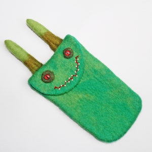 May include: A whimsical green felt case with a flap closure, featuring a stitched smile and red button eyes. Two green and brown felt horns extend from the top. The case is designed to hold small items.