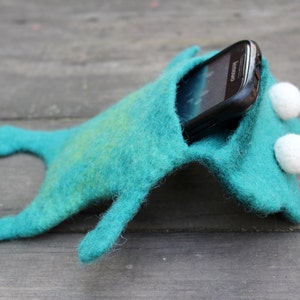 May include: A teal felt phone case shaped like a whimsical creature, with white, button-like eyes. A black smartphone is partially visible inside the case. The case has a playful, handmade aesthetic.