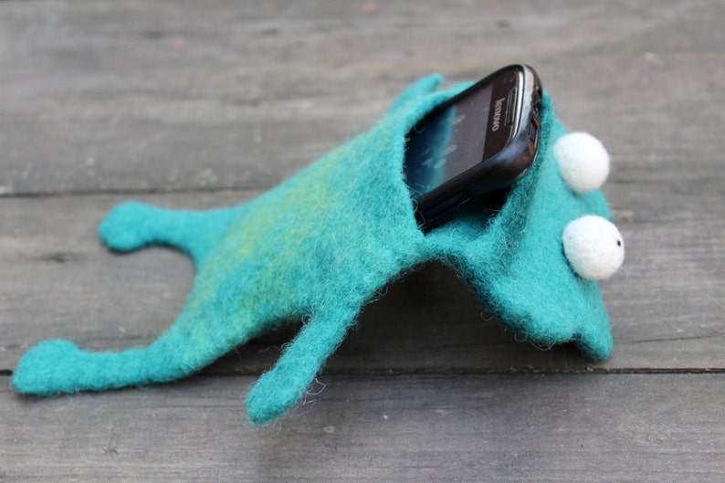 May include: A teal felt phone case shaped like a whimsical creature, with white, button-like eyes. A black smartphone is partially visible inside the case. The case has a playful, handmade aesthetic.