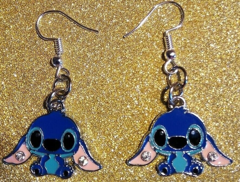 Puede incluir: Un par de pendientes de esmalte azul con un personaje de dibujos animados con ojos negros grandes, orejas rosas y vientre turquesa. Los pendientes tienen ganchos plateados y peque&ntilde;os detalles de cristal.
