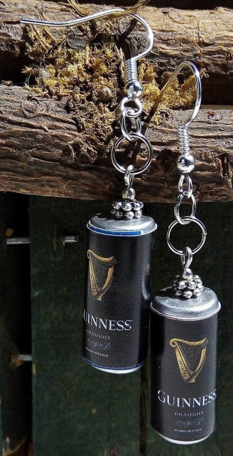May include: A pair of silver dangle earrings featuring miniature black Guinness cans with the iconic harp logo and the text "Guinness Draught Brewed in Dublin".
