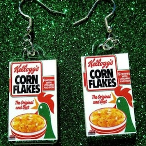 Op de afbeelding: Een paar oorbellen met miniatuurdoosjes Kellogg's Corn Flakes ontbijtgranen. De doosjes zijn rood, wit en groen met het Kellogg's logo en de tekst "Corn Flakes" in vetgedrukte, zwarte letters. De tekst "The Original and Best" is onder de naam van de ontbijtgranen gedrukt. De doosjes zijn bevestigd aan zilveren oorhaken.
