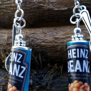 Könnte beinhalten: Ein Paar silberne Ohrringe mit einem Heinz Baked Beans Dosen-Anhänger. Die Anhänger sind blau mit einem weißen Etikett und roten Bohnen. Der Text auf dem Etikett lautet "Heinz Beans".