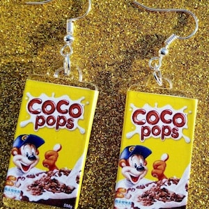 Puede incluir: Un par de pendientes con una caja amarilla con la mascota de cereales Coco Pops y el texto "Coco Pops" y "550g".