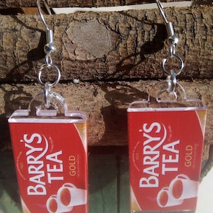 Op de afbeelding: Een paar rode en gouden oorbellen met het Barry's Tea logo. De oorbellen zijn gemaakt van helder acryl en hebben zilveren haakjes.