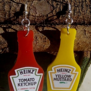Puede incluir: Un par de pendientes originales con forma de botellas de ketchup y mostaza amarilla Heinz. El pendiente rojo de ketchup tiene el texto "TOMATO KETCHUP" y el pendiente de mostaza amarilla tiene el texto "YELLOW MUSTARD".