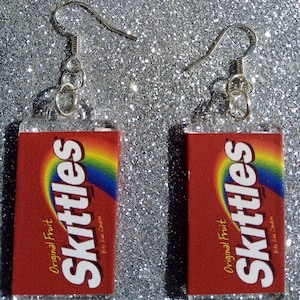 Puede incluir: Un par de pendientes de caramelo Skittles rojo y blanco. Los pendientes están hechos de plástico transparente y presentan un diseño de bolsa de caramelo Skittles con el texto "Original Fruit" y "Bite Size Candies".