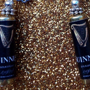 May include: A pair of silver earrings with black and gold Guinness beer bottle charms. The charms feature the iconic Guinness harp logo and the words "Guinness Draught" printed on the front.