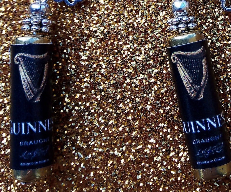 May include: A pair of silver earrings with black and gold Guinness beer bottle charms. The charms feature the iconic Guinness harp logo and the words "Guinness Draught" printed on the front.