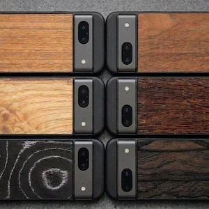 Op de afbeelding: Zes smartphonehoesjes gerangschikt in twee kolommen, elk met een zwarte camerabalk en een unieke houten inleg. De houttexturen variëren van lichtbruin tot donkerbruin en zwart, waarbij één hoesje een opvallend cirkelvormig houtnerfpatroon heeft.