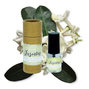 May include: A brown cardboard tube with a white label that says "Jasmine 100% Natural Phone Case Scent Refresher www.mmmore.net" and a clear glass bottle with a black cap and a white label that says "Jasmine 100% Natural Phone Case Scent Refresher www.mmmore.net". The bottle and tube are surrounded by white jasmine flowers and green leaves.