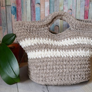 May include: A beige crocheted tote bag with white stripes. The bag has a single handle and is made of a soft, textured yarn.