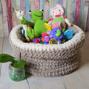 May include: A brown crocheted storage basket filled with a variety of toys, including a green frog, a pink doll, a giraffe, and a purple elephant. The basket is sitting on a wooden floor.