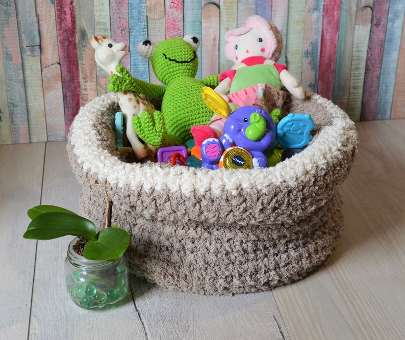 May include: A brown crocheted storage basket filled with a variety of toys, including a green frog, a pink doll, a giraffe, and a purple elephant. The basket is sitting on a wooden floor.