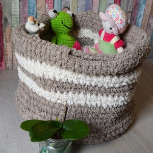 May include: A brown and white striped crocheted storage basket filled with stuffed toys, including a green frog, a white giraffe, and a pink and white bunny wearing a green dress. The basket is sitting on a light wood floor.