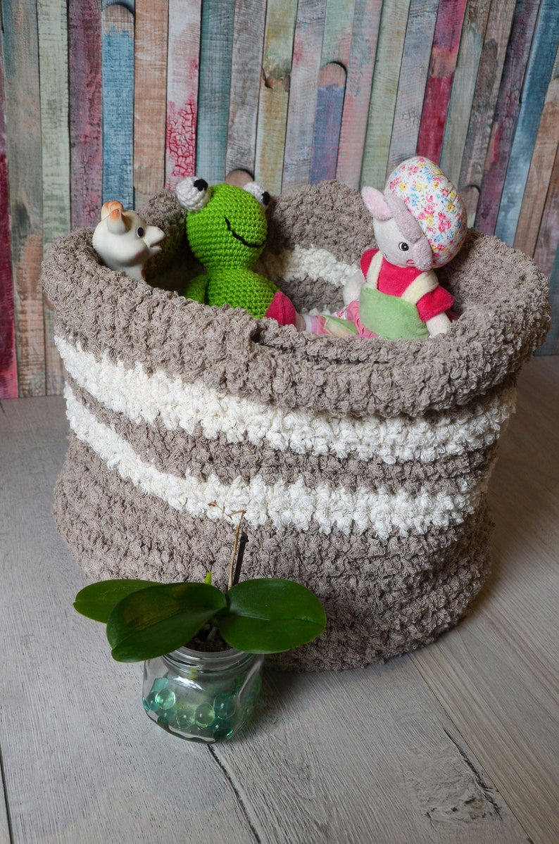 May include: A brown and white striped crocheted storage basket filled with stuffed toys, including a green frog, a white giraffe, and a pink and white bunny wearing a green dress. The basket is sitting on a light wood floor.