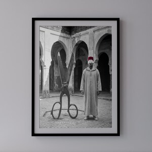 Puede incluir: Una fotografía enmarcada en blanco y negro que muestra a una persona con una chilaba clara y un fez rojo, con los ojos vendados, de pie junto a unas grandes tijeras de metal en un patio con arcos. La imagen destaca un marcado contraste.