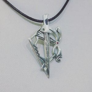 Può includere: Collana con pendente in argento con un design a spada, arco e freccia. Il pendente ha un piccolo accento rosso ed è appeso a un cordino nero. Il pendente è lungo circa 3,8 cm.