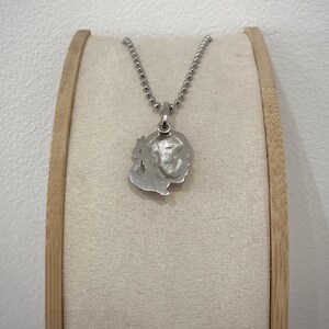 Puede incluir: Un collar de plata con un colgante que presenta un dise&ntilde;o detallado y texturizado de una cara. El colgante est&aacute; unido a una cadena de plata.