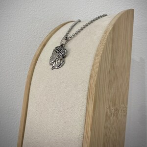 Puede incluir: Un collar de plata con un colgante detallado que presenta la cara de una mujer. El collar se muestra en un soporte de madera con una cubierta de tela beige.
