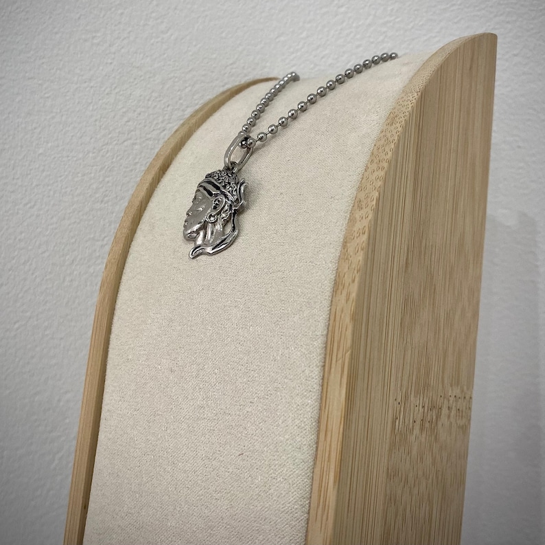 Puede incluir: Un collar de plata con un colgante detallado que presenta la cara de una mujer. El collar se muestra en un soporte de madera con una cubierta de tela beige.
