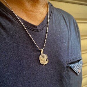 Puede incluir: Un collar de cadena de plata con un colgante de plata que presenta un perfil detallado de la cara de una persona. El colgante lo lleva una persona que lleva una camisa azul oscuro.