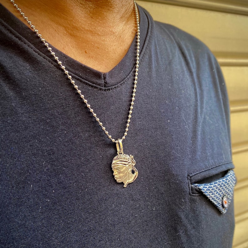 Puede incluir: Un collar de cadena de plata con un colgante de plata que presenta un perfil detallado de la cara de una persona. El colgante lo lleva una persona que lleva una camisa azul oscuro.