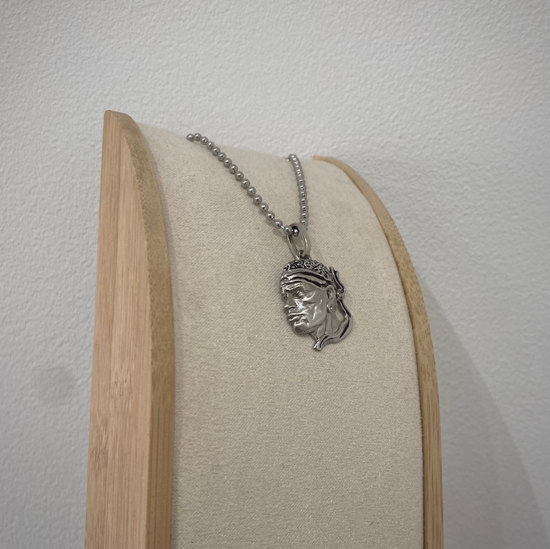 Puede incluir: Un collar de cadena de plata con un colgante de plata que presenta un perfil detallado de la cara de un hombre con un pa&ntilde;uelo.