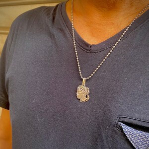 Puede incluir: Un collar de cadena de plata con un colgante de plata que presenta el perfil de una persona con un turbante.