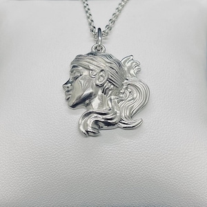 May include: A silver-colored pendant necklace featuring a profile of a woman's face with flowing hair. The pendant is attached to a delicate chain. The woman's profile is detailed, with closed eyes and a graceful neck.