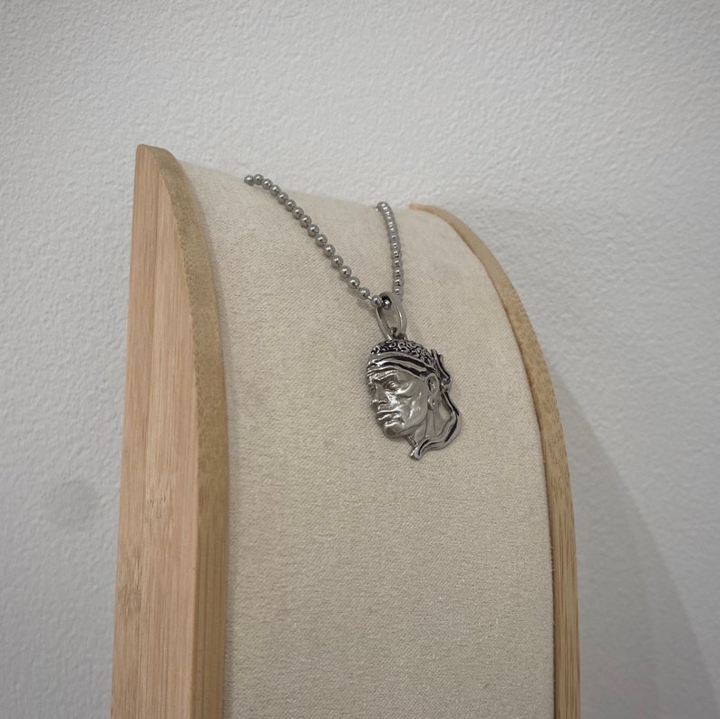 Puede incluir: Un collar de cadena de plata con un colgante que presenta un perfil detallado de un hombre con un pa&ntilde;uelo. El collar se muestra en un soporte de madera.