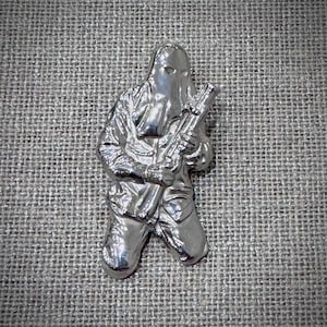 Puede incluir: Una figura de metal plateado que representa a una persona arrodillada sosteniendo un rifle. La figura lleva una capucha y ropa de estilo militar. Sobre una superficie gris texturizada.