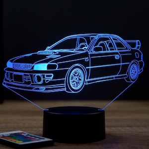 Op de afbeelding: Een blauw LED-licht met een 3D-omtrek van een Subaru Impreza-auto. Het licht staat op een zwarte basis en heeft een afstandsbediening.