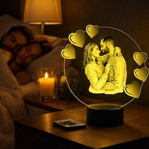 Könnte beinhalten: Ein personalisiertes Nachtlicht mit einem sich umarmenden Paar, umgeben von herzförmigen Akzenten. Die Lampe strahlt ein warmes gelbes Licht aus, mit einem schwarzen Sockel. Eine Fernbedienung liegt in der Nähe. Eine Kerze und ein schlafendes Paar sind im Hintergrund.