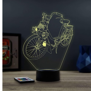 Könnte beinhalten: Eine LED-Lampe in Motorradform mit schwarzem Sockel. Die Lampe strahlt ein sanftes gelbes Licht aus, das das Design des Motorrads hervorhebt. Eine Fernbedienung liegt auf der Holzoberfläche vor der Lampe. Der Hintergrund ist dunkelgrau.