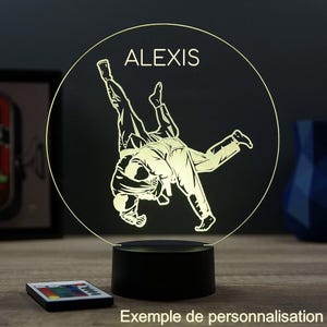 Peut inclure: Une veilleuse LED personnalisée avec une illustration de judo. Le design comprend le nom "ALEXIS" en lettres blanches au-dessus des figures de judo. La base est noire, et une télécommande est visible.