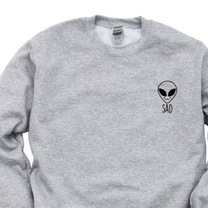 Puede incluir: Una sudadera gris con una pequeña cara de extraterrestre negra y la palabra "SAD" impresa en la parte delantera.