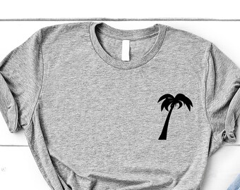 Könnte beinhalten: Ein graues T-Shirt mit einem schwarzen Palmenbaum-Motiv auf der linken Brust. Das T-Shirt liegt flach auf einer weißen Holzoberfläche mit einem Paar weißen Sneakers und einer hellblauen Jeans.